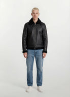 Eine Person mit kurzen blonden Haaren schaut nach vorne und trägt die trueprodigy Herren Lederjacke aus Schafsleder in Schwarz im Aviator-Stil mit Fellbesatz, gepaart mit hellblauen Jeans und weißen Turnschuhen vor einem schlichten hellen Hintergrund.