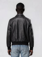 Man sieht eine Person von hinten, die die trueprodigy Herren Lederjacke aus Schafsleder in Schwarz mit klassischem Blouson-Schnitt und hellblaue Jeans vor einem schlichten, hellen Hintergrund trägt.