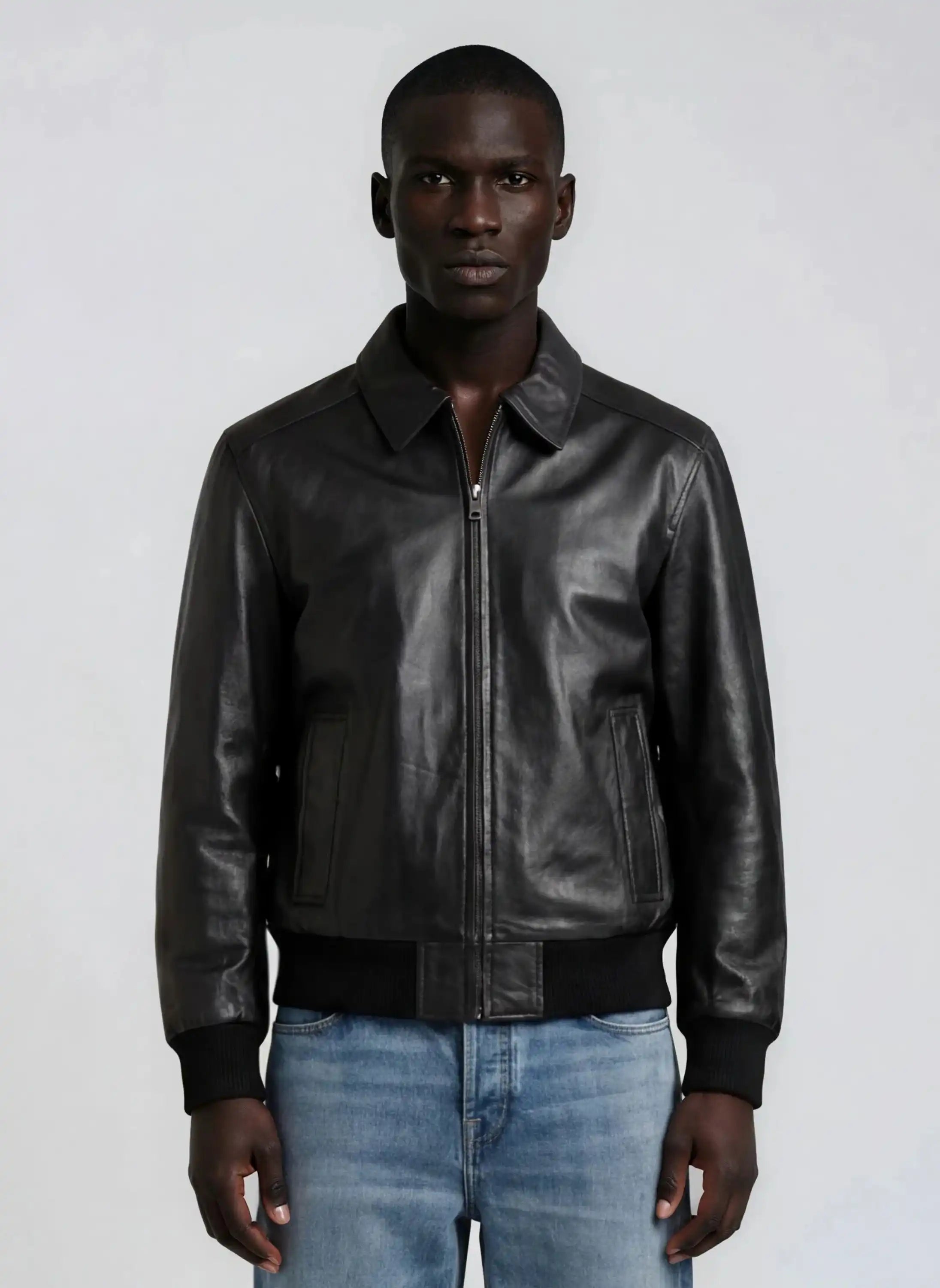 A man in a trueprodigy Herren Lederjacke aus Schafsleder in Schwarz mit klassischem Blouson-Schnitt, über einem hellen Shirt und blauen Jeans, steht vor hellem Hintergrund und blickt in die Kamera.