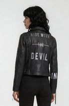 Eine Person mit langem, welligem schwarzem Haar trägt eine trueprodigy Bikerlederjacke mit silbernen Nieten, weißem "RIDE WITH THE DEVIL"-Aufdruck und dazu passenden schwarzen Hosen.