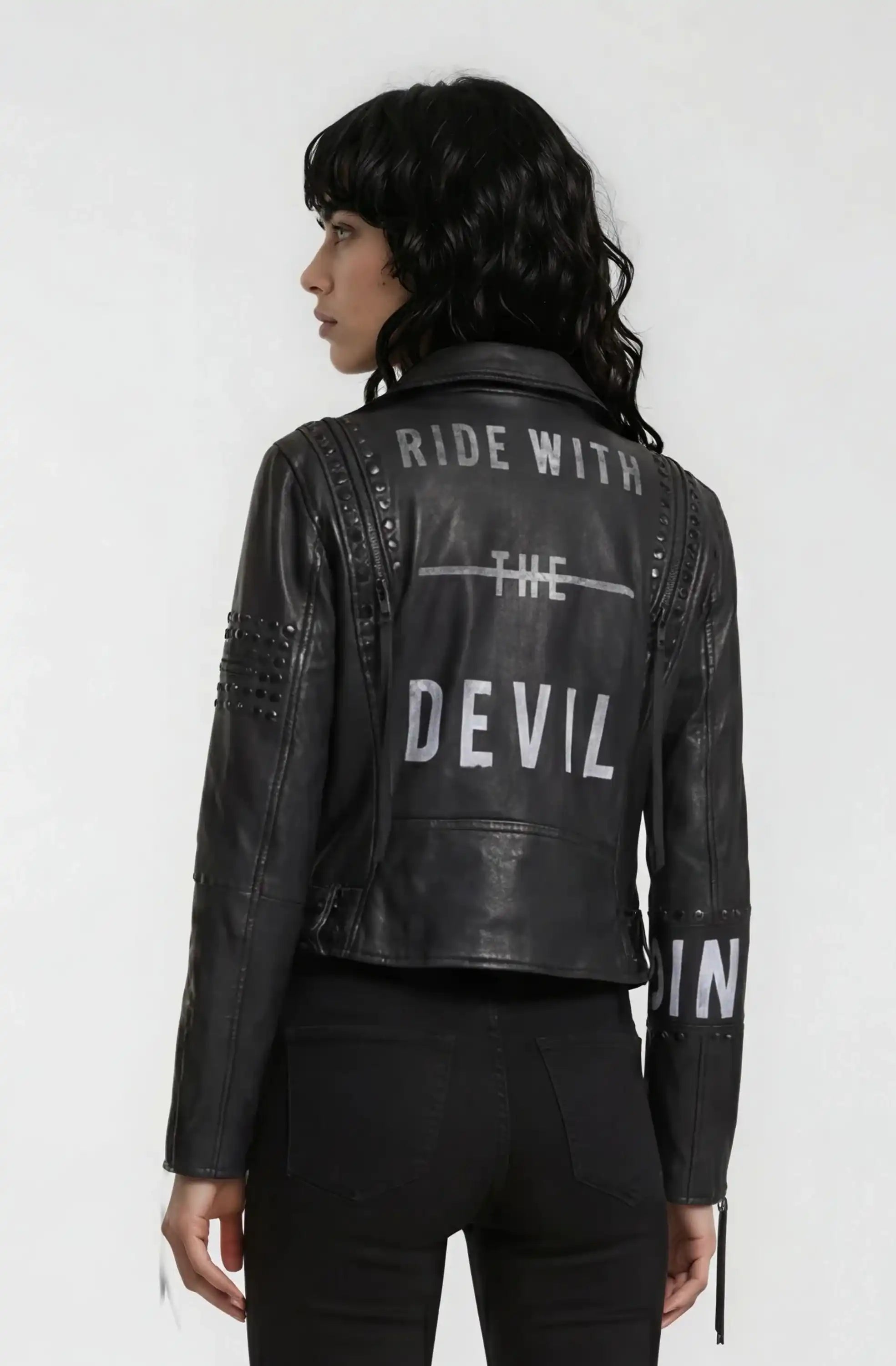 Eine Person mit langem, welligem schwarzem Haar trägt eine trueprodigy Bikerlederjacke mit silbernen Nieten, weißem "RIDE WITH THE DEVIL"-Aufdruck und dazu passenden schwarzen Hosen.