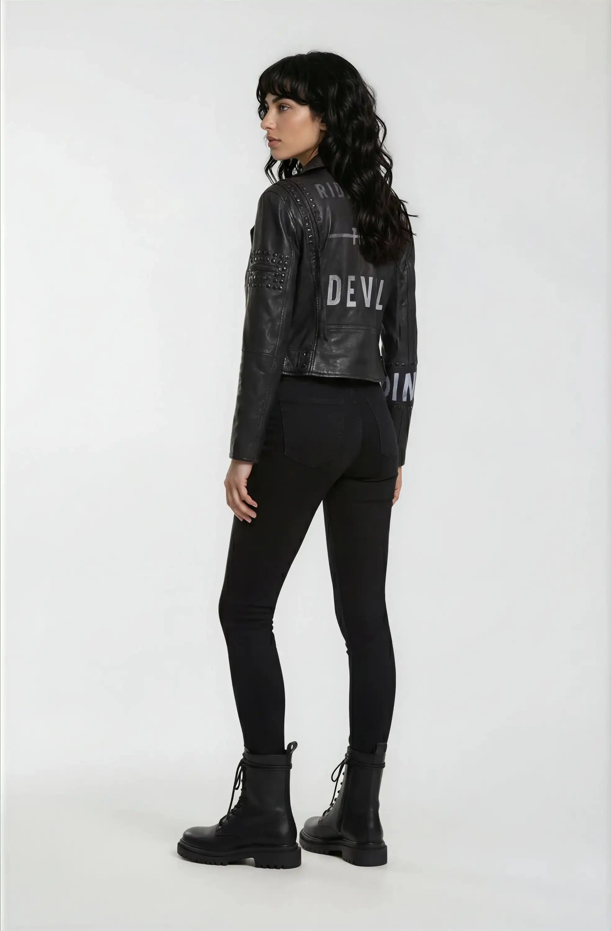 Eine Person mit langen dunklen Haaren, bekleidet mit der trueprodigy Bikerlederjacke mit Nieten und Print, schwarzen Skinny-Jeans und schwarzen Kampfstiefeln steht mit dem Gesicht zur Kamera vor einem schlichten weißen Hintergrund.