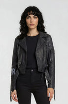 Eine Frau mit welligem, dunklem Haar trägt die Bikerlederjacke mit Nieten und Print von trueprodigy über schwarzem Shirt und Hose, stehend vor einem schlichten, hellgrauen Hintergrund.