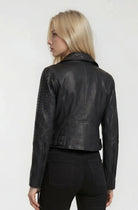 Eine Person mit langen blonden Haaren, bekleidet mit einer schwarzen Trueprodigy Bikerlederjacke mit Ösen und einer schwarzen Hose, steht von der Kamera abgewandt vor einem schlichten hellen Hintergrund.