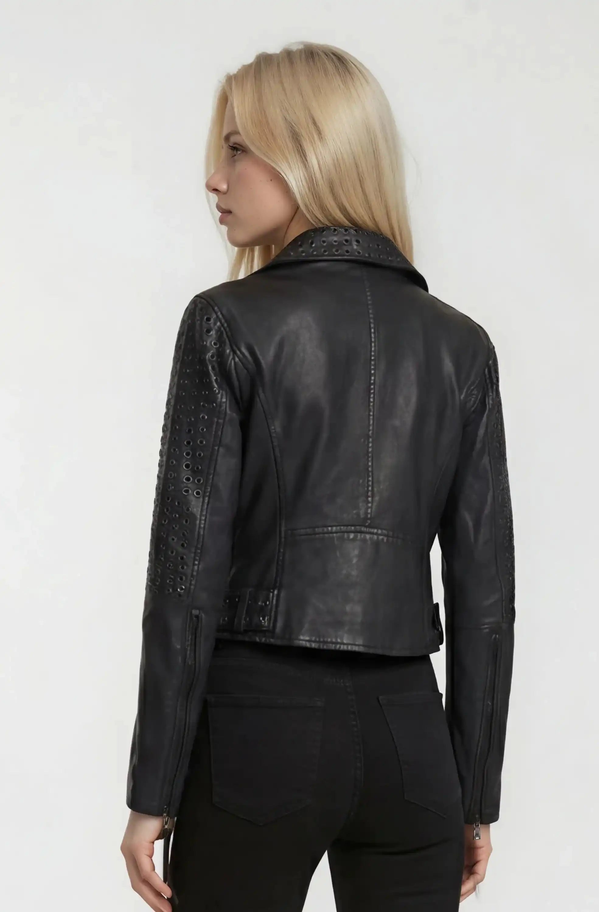 Eine Person mit langen blonden Haaren, bekleidet mit einer schwarzen Trueprodigy Bikerlederjacke mit Ösen und einer schwarzen Hose, steht von der Kamera abgewandt vor einem schlichten hellen Hintergrund.