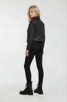 Eine Frau mit langen blonden Haaren blickt über ihre Schulter. Sie trägt eine schwarze Trueprodigy Bikerlederjacke mit Ösen, eine schwarze Hose und schwarze Schnürstiefel vor einem schlichten weißen Hintergrund.