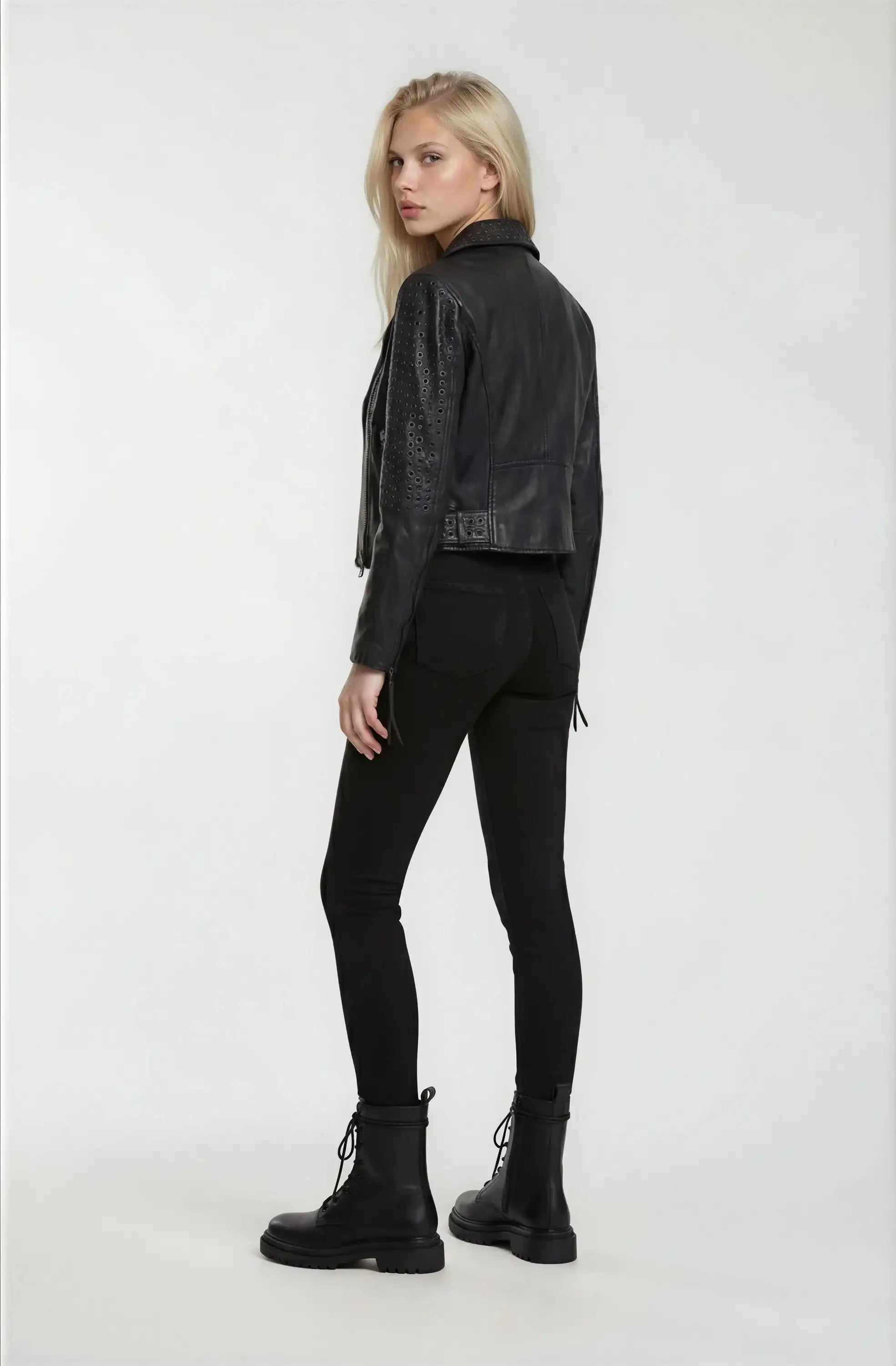 Eine Frau mit langen blonden Haaren blickt über ihre Schulter. Sie trägt eine schwarze Trueprodigy Bikerlederjacke mit Ösen, eine schwarze Hose und schwarze Schnürstiefel vor einem schlichten weißen Hintergrund.