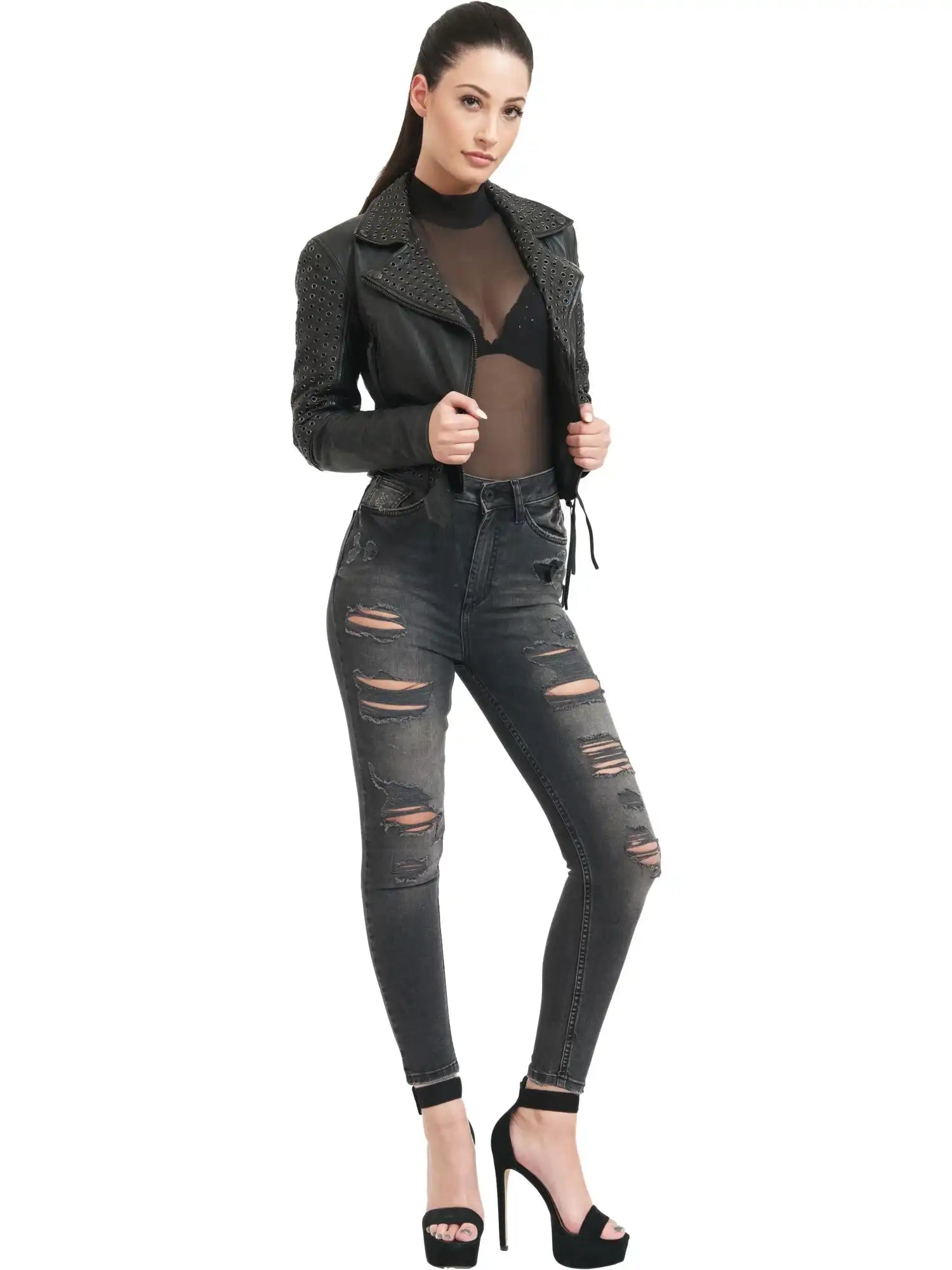 Eine Frau steht vor einem weißen Hintergrund und trägt eine schwarze Trueprodigy Raven Bikerlederjacke mit Ösen, ein durchsichtiges schwarzes Oberteil mit sichtbarem schwarzem BH, ausgebeulte Skinny Jeans und schwarze hochhackige Sandalen.