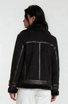 Eine Person mit langen dunklen Haaren trägt eine schwarze Trueprodigy Wilderlederjacke mit Gürtel und eine weiße Hose und blickt von der Kamera weg vor einem schlichten hellen Hintergrund.