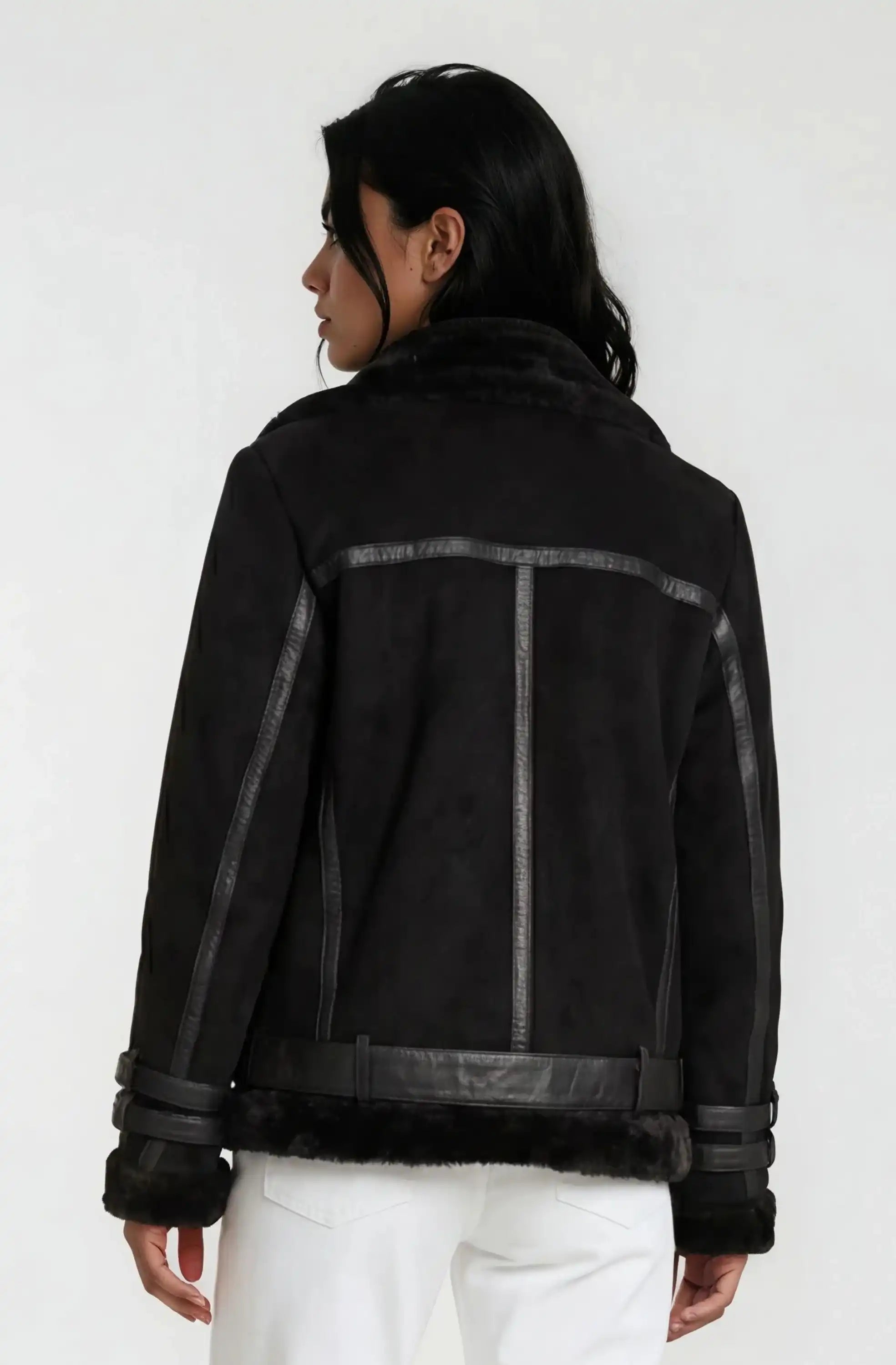 Eine Person mit langen dunklen Haaren trägt eine schwarze Trueprodigy Wilderlederjacke mit Gürtel und eine weiße Hose und blickt von der Kamera weg vor einem schlichten hellen Hintergrund.