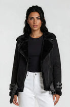 Eine Person mit langen dunklen Haaren trägt eine Trueprodigy Wilderlederjacke mit Gürtel über einem schwarzen Oberteil und einer weißen Hose mit hoher Taille vor einem schlichten hellen Hintergrund.