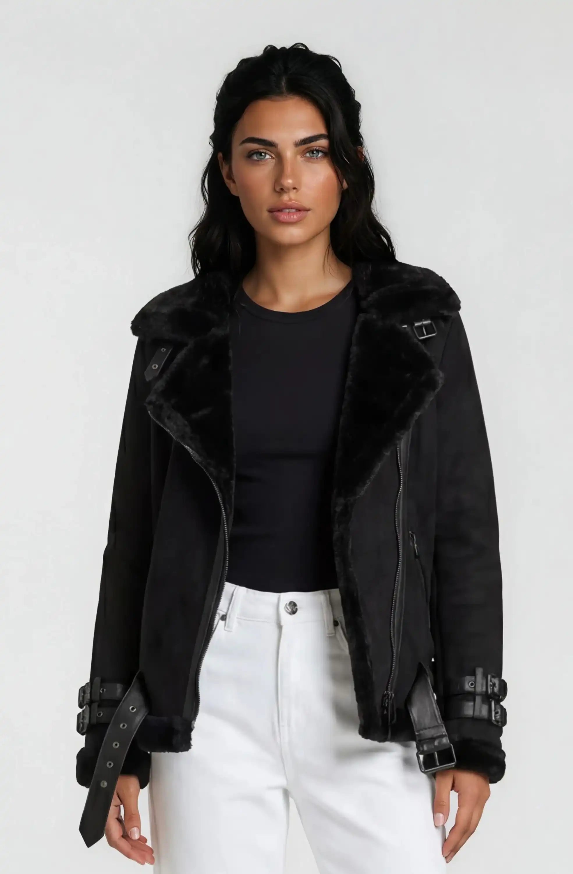 Eine Person mit langen dunklen Haaren trägt eine Trueprodigy Wilderlederjacke mit Gürtel über einem schwarzen Oberteil und einer weißen Hose mit hoher Taille vor einem schlichten hellen Hintergrund.