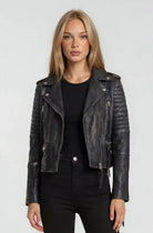 Eine Frau mit glattem blondem Haar trägt die trueprodigy Bikerlederjacke Vintage Optik über einem schwarzen Oberteil und einer Hose, steht vor einem schlichten hellgrauen Hintergrund und schaut direkt in die Kamera.