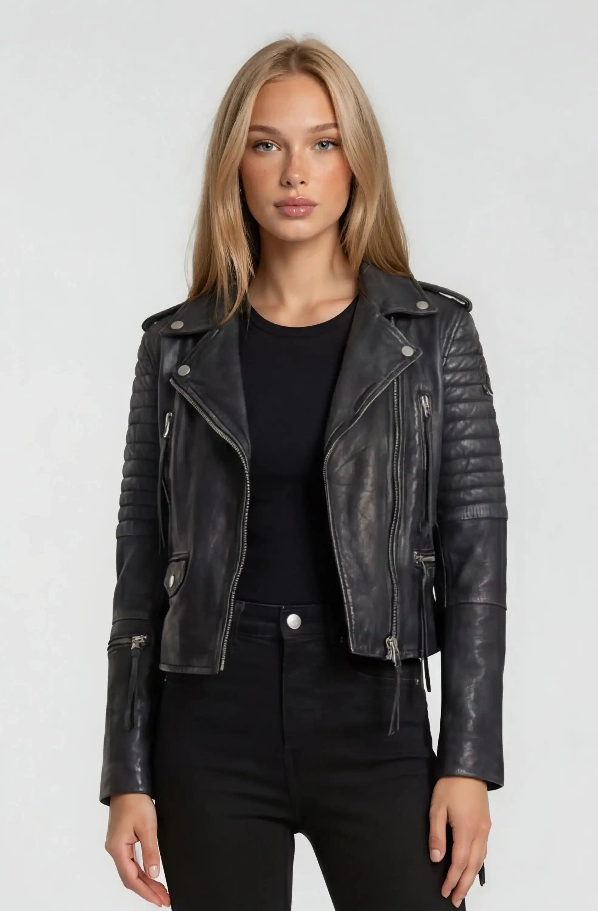 Eine Frau mit glattem blondem Haar trägt die trueprodigy Bikerlederjacke Vintage Optik über einem schwarzen Oberteil und einer Hose, steht vor einem schlichten hellgrauen Hintergrund und schaut direkt in die Kamera.