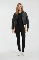 Eine Frau mit glattem blondem Haar und neutralem Gesichtsausdruck blickt nach vorne. Sie trägt die trueprodigy Bikerlederjacke Vintage Optik in Schwarz, ein schwarzes Hemd, High-Waisted Skinny Jeans und weiße Turnschuhe vor einem schlichten hellen Hintergrund.