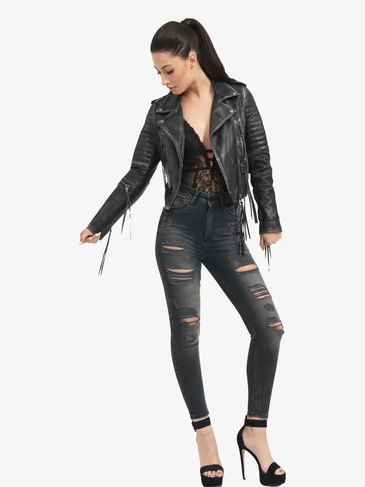 Eine Frau mit langen dunklen Haaren in einem Pferdeschwanz trägt die Avril Bikerlederjacke Vintage Optik von trueprodigy in Schwarz, gepaart mit einem schwarzen Spitzentop, zerrissenen dunklen Skinny Jeans und schwarzen hochhackigen Sandalen, vor einem schlichten weißen Hintergrund.