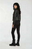 Eine Person mit langen, gewellten dunklen Haaren trägt die trueprodigy Biker Lederjacke, eine schwarze Hose und schwarze Stiefel. Sie steht und schaut über ihre Schulter vor einem schlichten weißen Hintergrund.