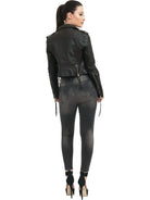 Eine Frau mit Pferdeschwanz steht mit dem Gesicht zur Wand und trägt die trueprodigy Jade Biker Lederjacke, verwaschene schwarze Skinny Jeans und schwarze hochhackige Schuhe vor einem schlichten weißen Hintergrund.