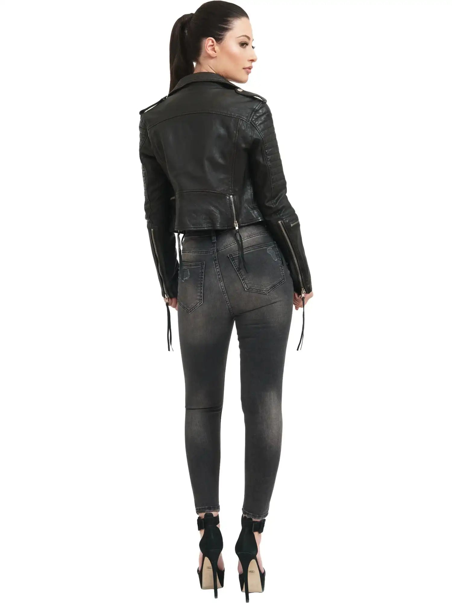 Eine Frau mit Pferdeschwanz steht mit dem Gesicht zur Wand und trägt die trueprodigy Jade Biker Lederjacke, verwaschene schwarze Skinny Jeans und schwarze hochhackige Schuhe vor einem schlichten weißen Hintergrund.
