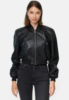 Eine Frau steht mit dem Blick nach vorne und trägt die trueprodigy Paola Kurze Oversized Lederjacke, eine schwarze Lederjacke mit Reißverschluss, gepaart mit einer schwarzen High-Waisted-Jeans vor einem schlichten hellen Hintergrund.