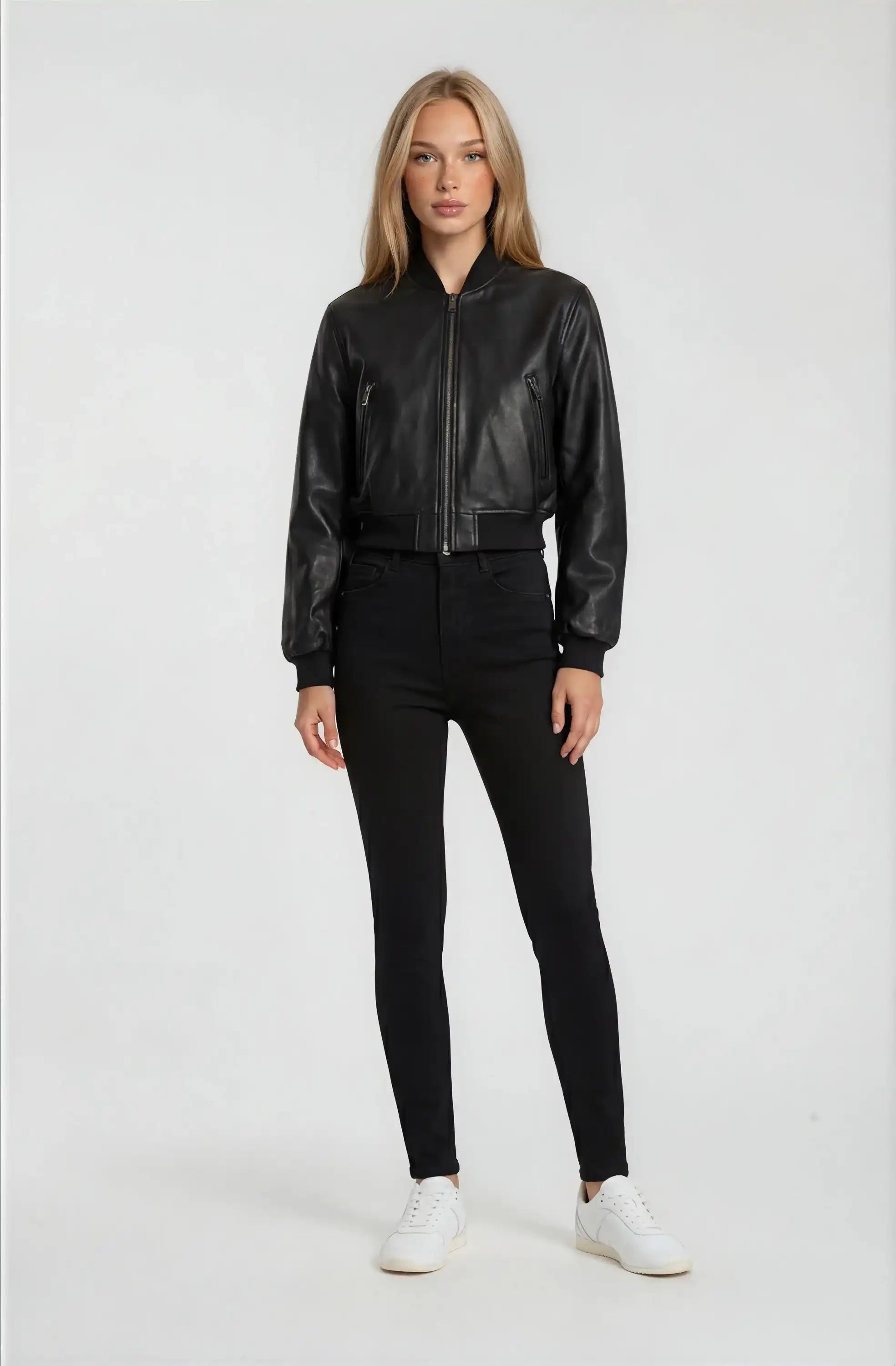 Eine Frau blickt vor einem schlichten weißen Hintergrund nach vorne und trägt eine schwarze Kurze Oversized Lederjacke von trueprodigy, eine schwarze Hose mit hoher Taille und weiße Turnschuhe. Ihre Arme sind entspannt an ihren Seiten.