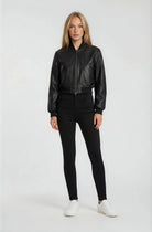 Eine Frau blickt vor einem schlichten weißen Hintergrund nach vorne und trägt eine schwarze Kurze Oversized Lederjacke von trueprodigy, eine schwarze Hose mit hoher Taille und weiße Turnschuhe. Ihre Arme sind entspannt an ihren Seiten.