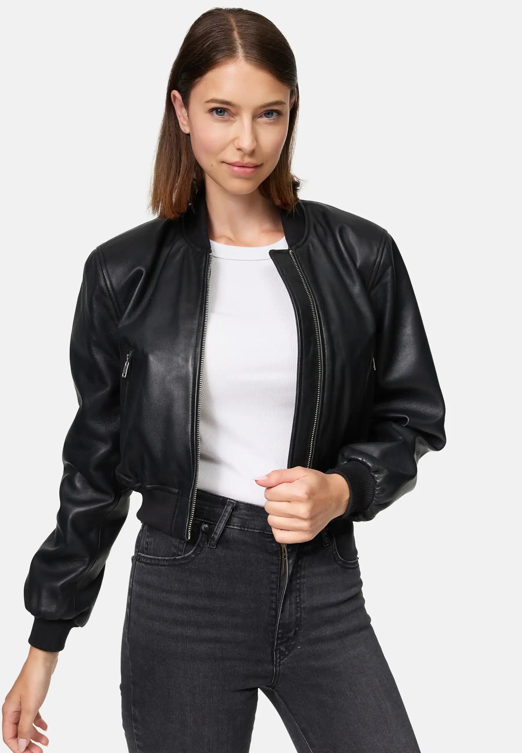 Eine Frau trägt die Paola Kurze Oversized Lederjacke von trueprodigy über einem weißen Shirt und dunklen Jeans. Sie steht vor einem schlichten Hintergrund, hat glattes braunes Haar und hält den Jackenreißverschluss mit einer Hand.