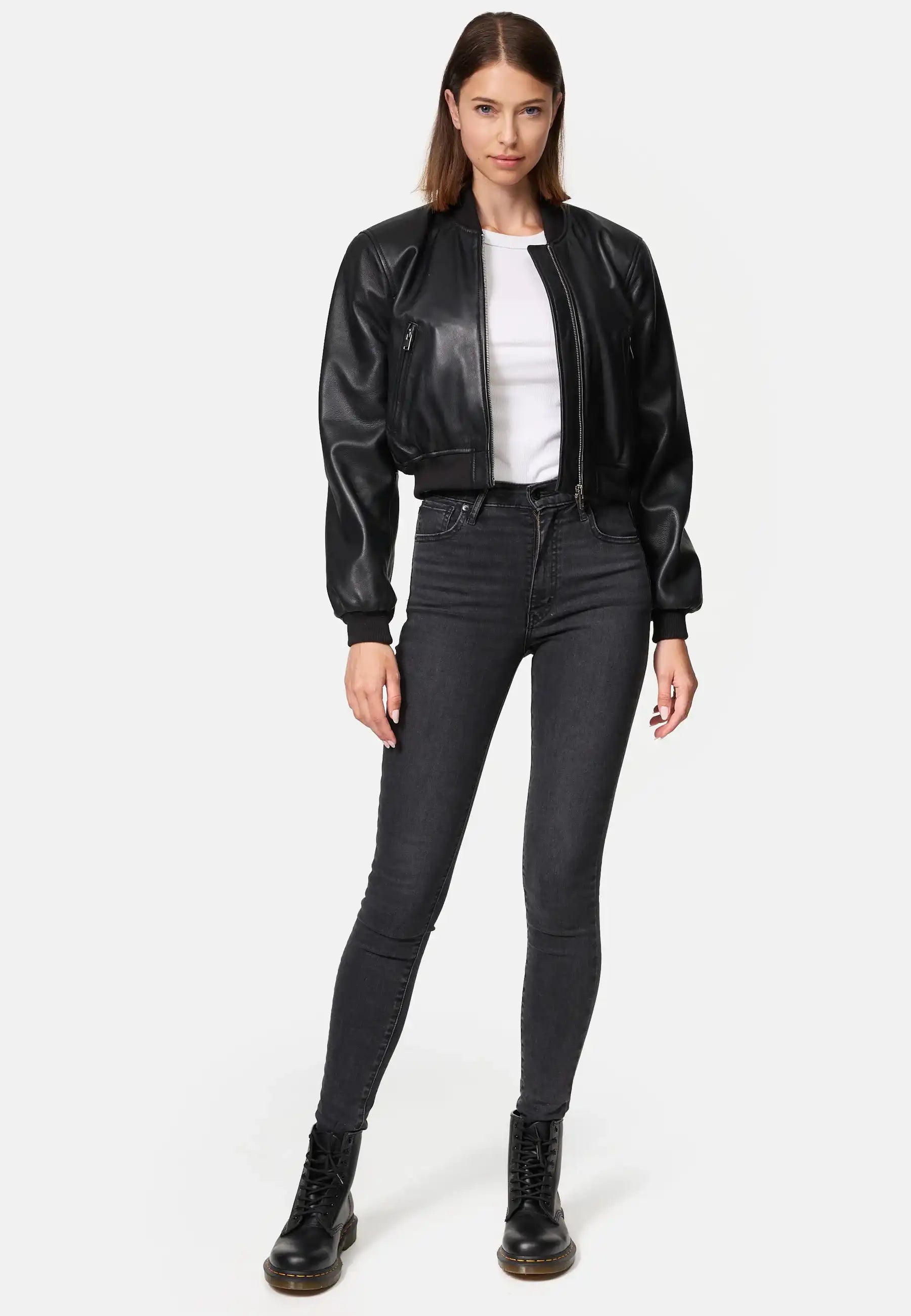 Eine Frau steht mit dem Gesicht nach vorne in einer Paola Kurze Oversized Lederjacke, einem weißen T-Shirt, einer schwarzen Jeans mit hoher Taille und schwarzen Schnürstiefeln vor einem schlichten weißen Hintergrund.