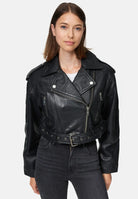 Eine Frau mit glattem braunem Haar trägt eine Trueprodigy Rosa Kurze Bomber-Lederjacke über einem weißen Oberteil und einer schwarzen Jeans mit hoher Taille. Sie steht vor einem schlichten hellen Hintergrund.