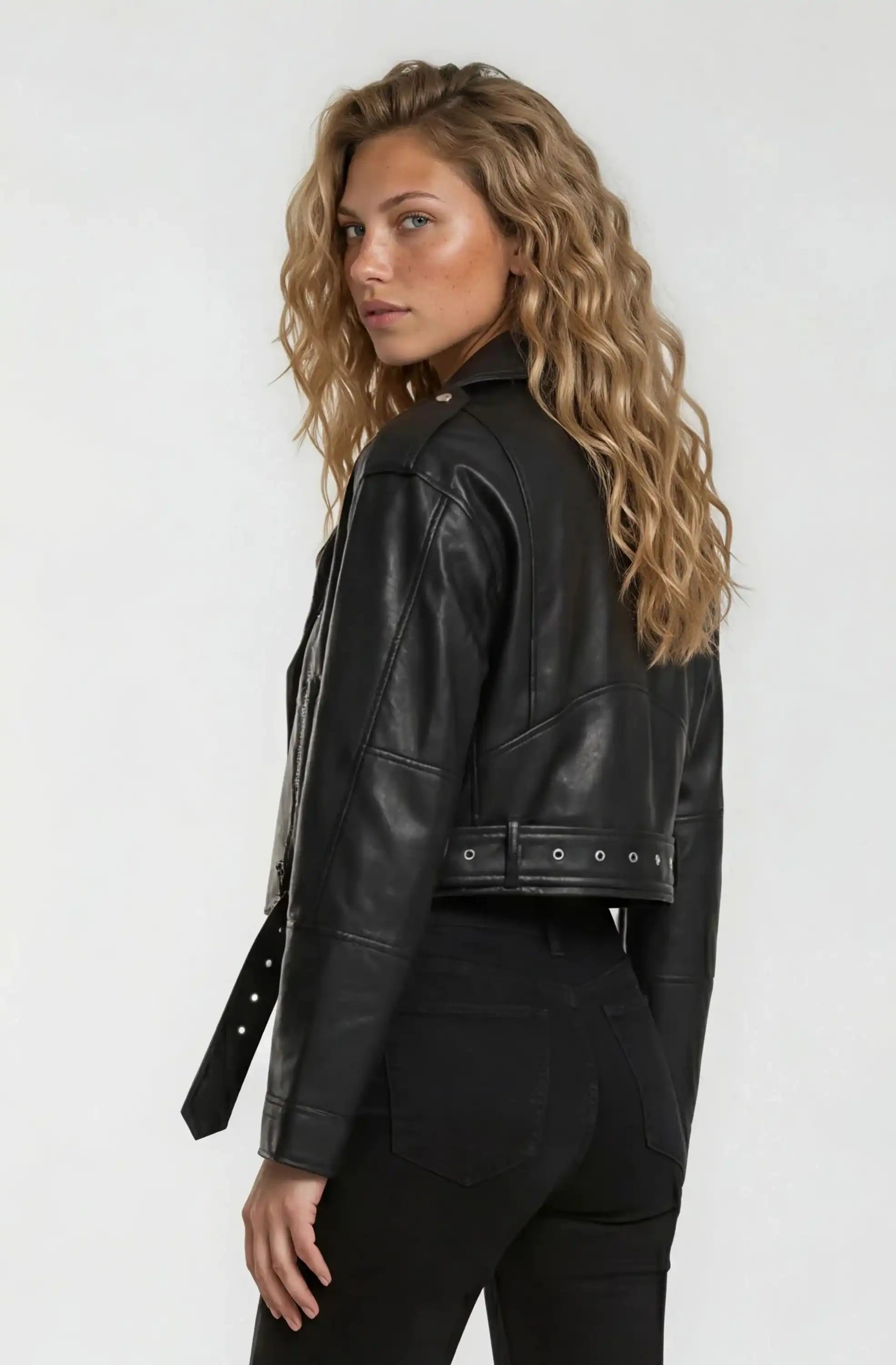 Eine Person mit langen, gewellten blonden Haaren trägt eine trueprodigy Kurze Bomber-Lederjacke und eine schwarze Hose, steht vor einem schlichten hellen Hintergrund und schaut über die Schulter in die Kamera.