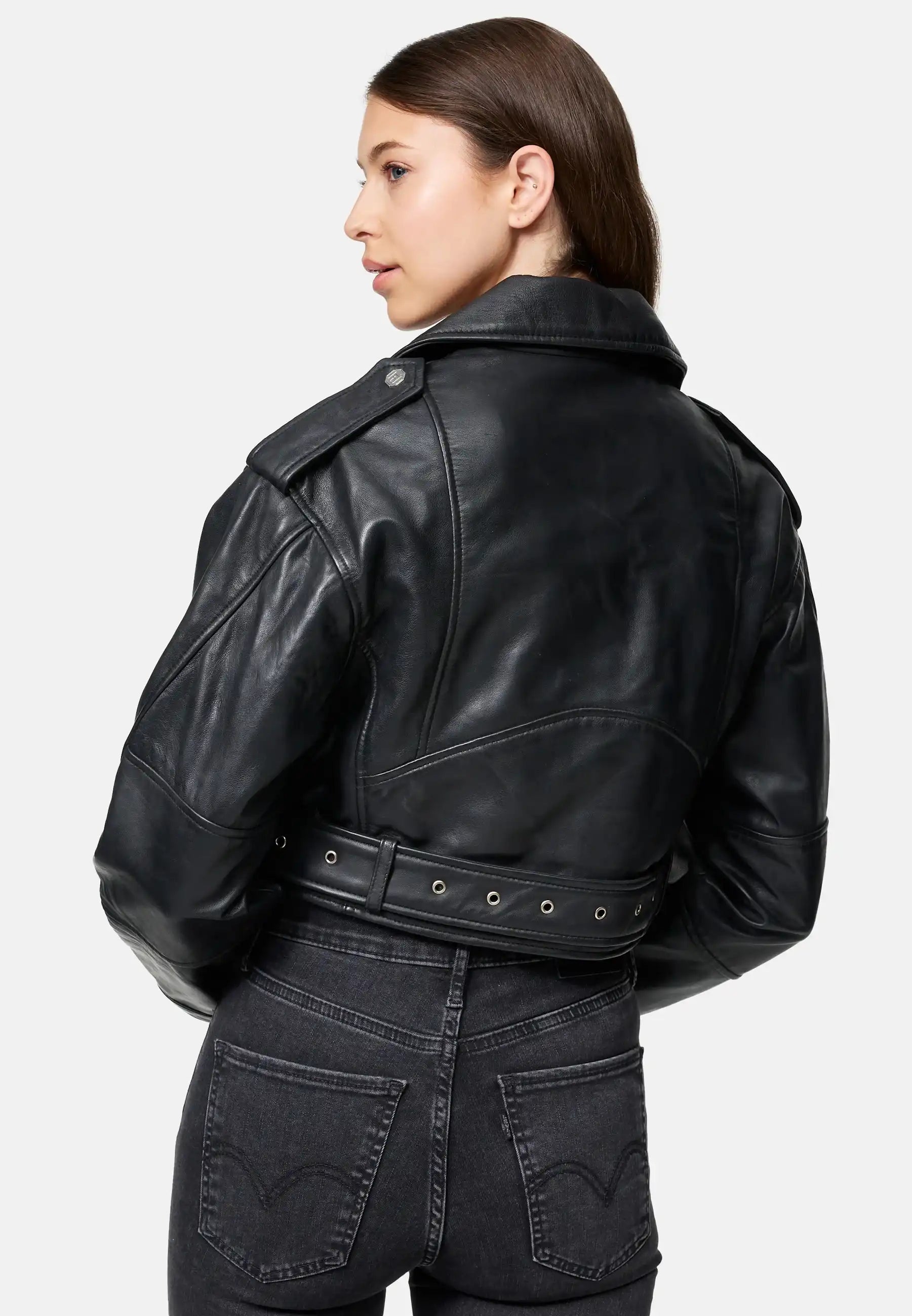 Eine Frau mit langen braunen Haaren trägt die trueprodigy Rosa Kurze Bomber-Lederjacke und schwarze Jeans. Sie steht mit dem Rücken zur Kamera und schaut zur Seite vor einem schlichten hellgrauen Hintergrund.