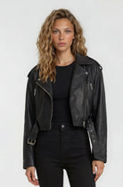 Eine Person mit gewelltem blondem Haar trägt eine trueprodigy Kurze Bomber-Lederjacke über einem schwarzen Hemd und einer schwarzen Hose und steht vor einem schlichten hellen Hintergrund.