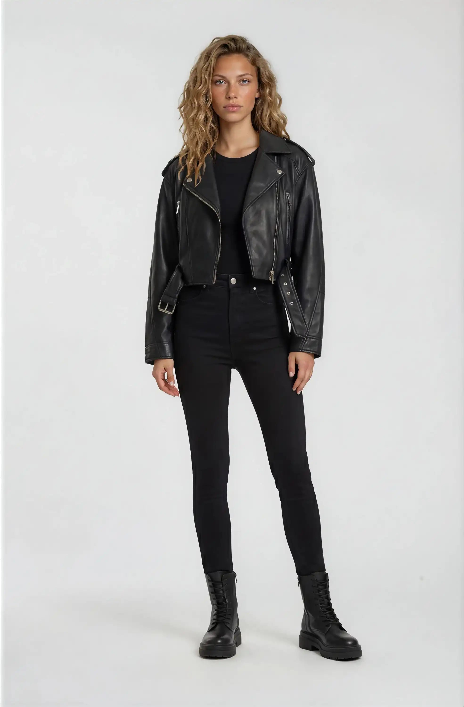 Eine Frau steht mit dem Gesicht nach vorne und trägt eine trueprodigy Kurze Bomber-Lederjacke im Biker-Stil, gepaart mit einem schwarzen T-Shirt, einer schwarzen Hose mit hoher Taille und schwarzen Schnürstiefeln vor einem schlichten hellen Hintergrund.
