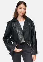 Eine Frau mit glattem braunem Haar trägt die Rosa Kurze Bomber-Lederjacke von trueprodigy über einem weißen Hemd und schwarzen Jeans. Sie steht vor einem schlichten weißen Hintergrund, eine Hand in der Tasche, die andere hält ihre Jacke.