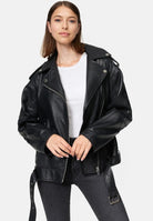 Eine Frau steht mit dem Gesicht nach vorne und trägt eine Trueprodigy Dua Lange übergroße Bikerlederjacke in Schwarz über einem weißen T-Shirt und dunkelgrauen Jeans. Sie hat glattes, schulterlanges braunes Haar und hält die Vorderseite der Jacke mit beiden Händen fest.