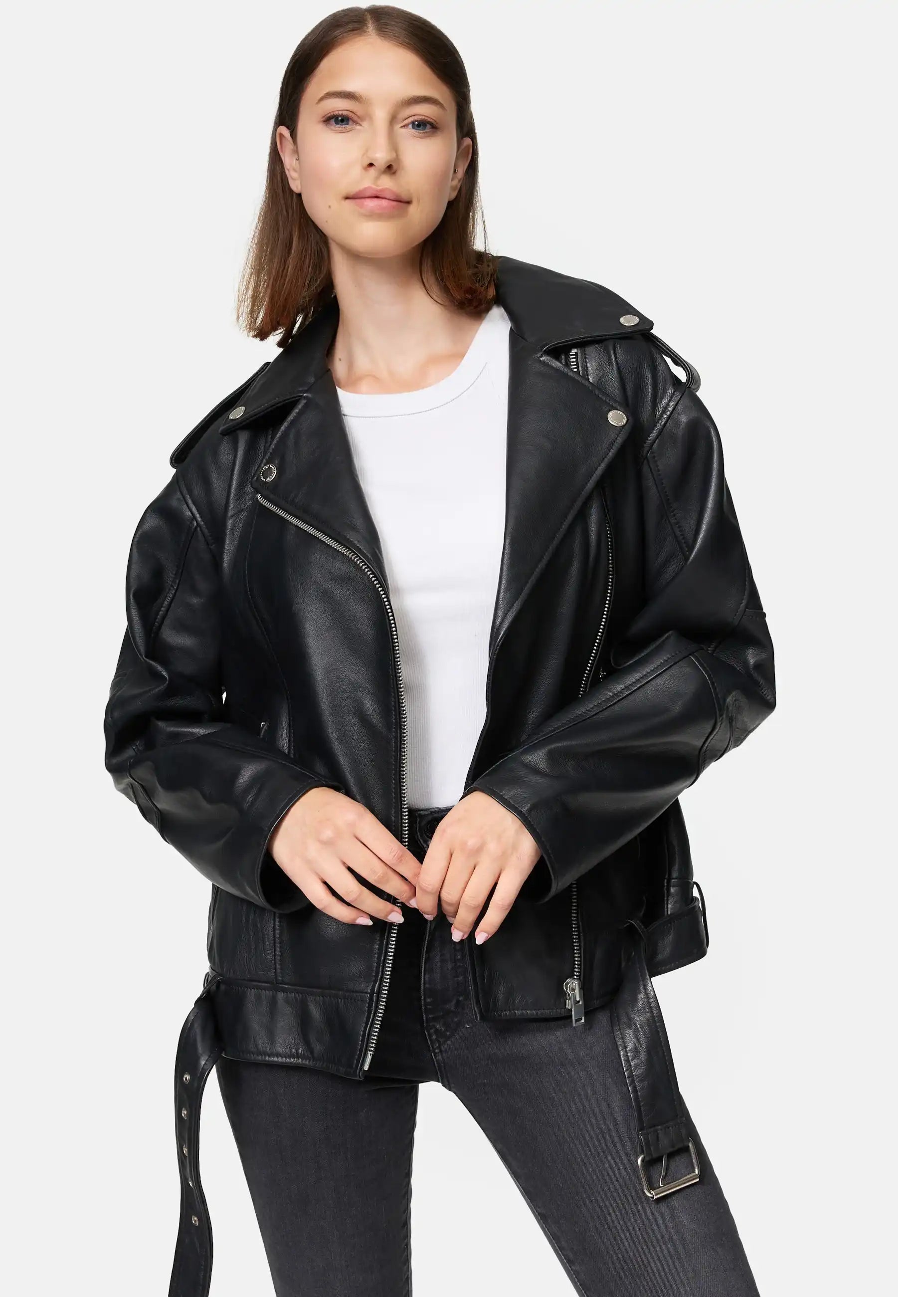 Eine Frau steht mit dem Gesicht nach vorne und trägt eine Trueprodigy Dua Lange übergroße Bikerlederjacke in Schwarz über einem weißen T-Shirt und dunkelgrauen Jeans. Sie hat glattes, schulterlanges braunes Haar und hält die Vorderseite der Jacke mit beiden Händen fest.