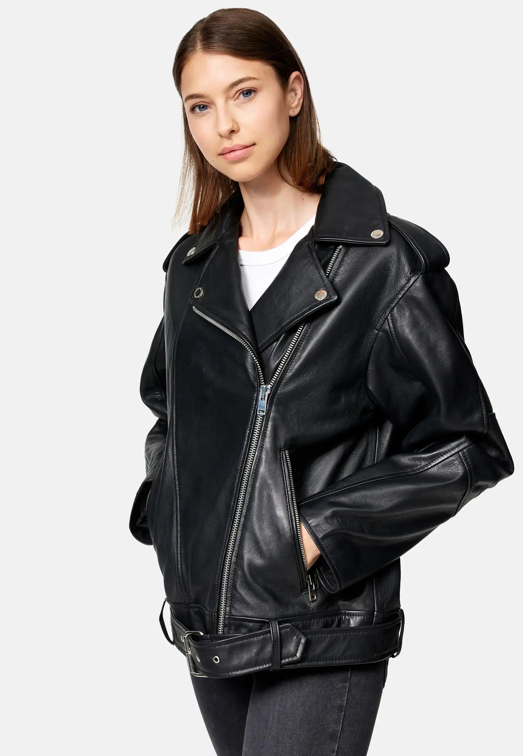 Eine Frau trägt eine trueprodigy Dua Lange übergroße Bikerlederjacke in Schwarz über einem weißen Oberteil und dunklen Jeans und steht mit den Händen in den Jackentaschen vor einem schlichten hellgrauen Hintergrund.