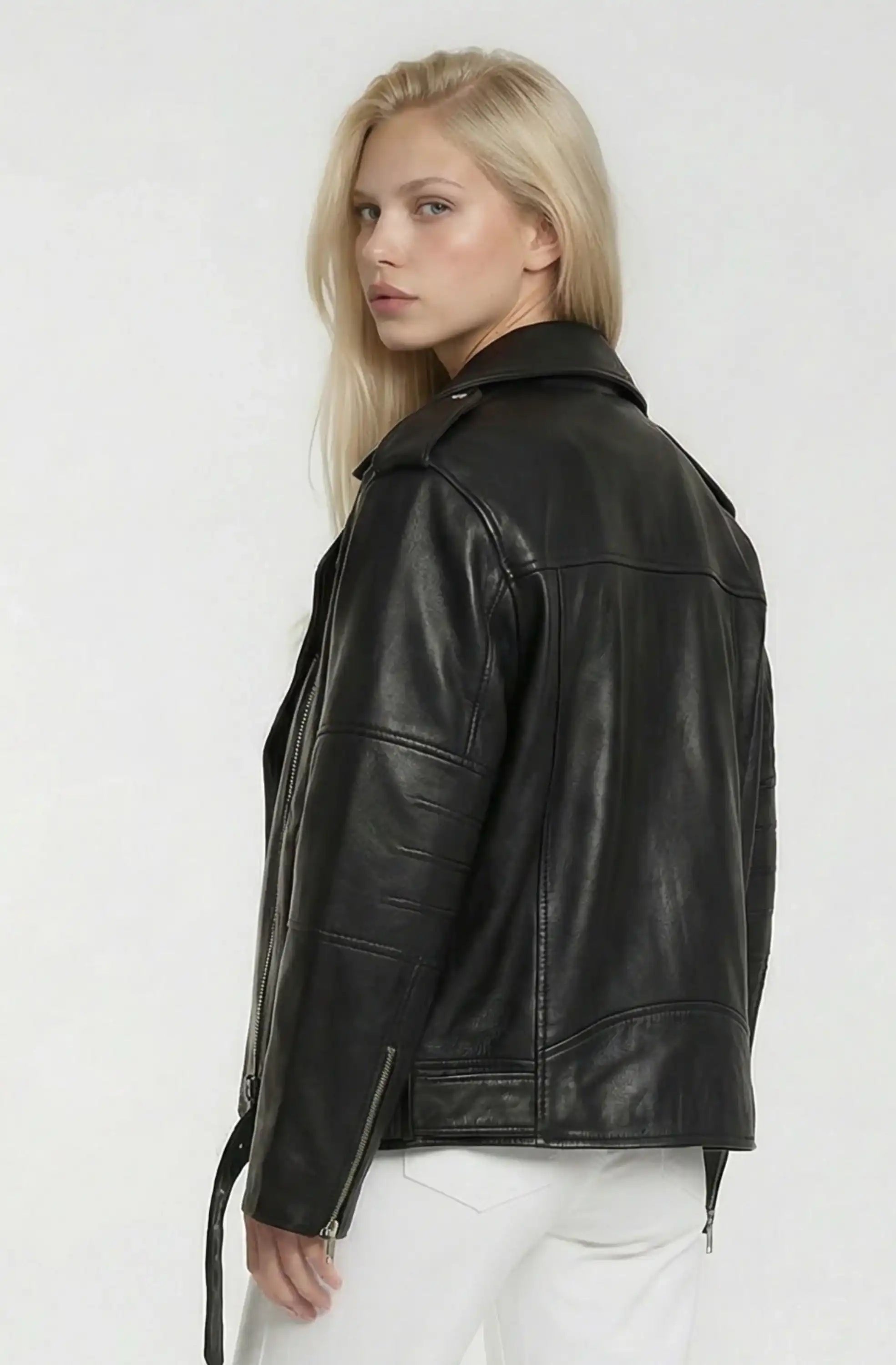 Eine Person mit langen blonden Haaren modelliert die trueprodigy Lange übergroße Bikerlederjacke und weiße Hose, steht vor einem schlichten hellen Hintergrund und schaut über die Schulter in Richtung Kamera.