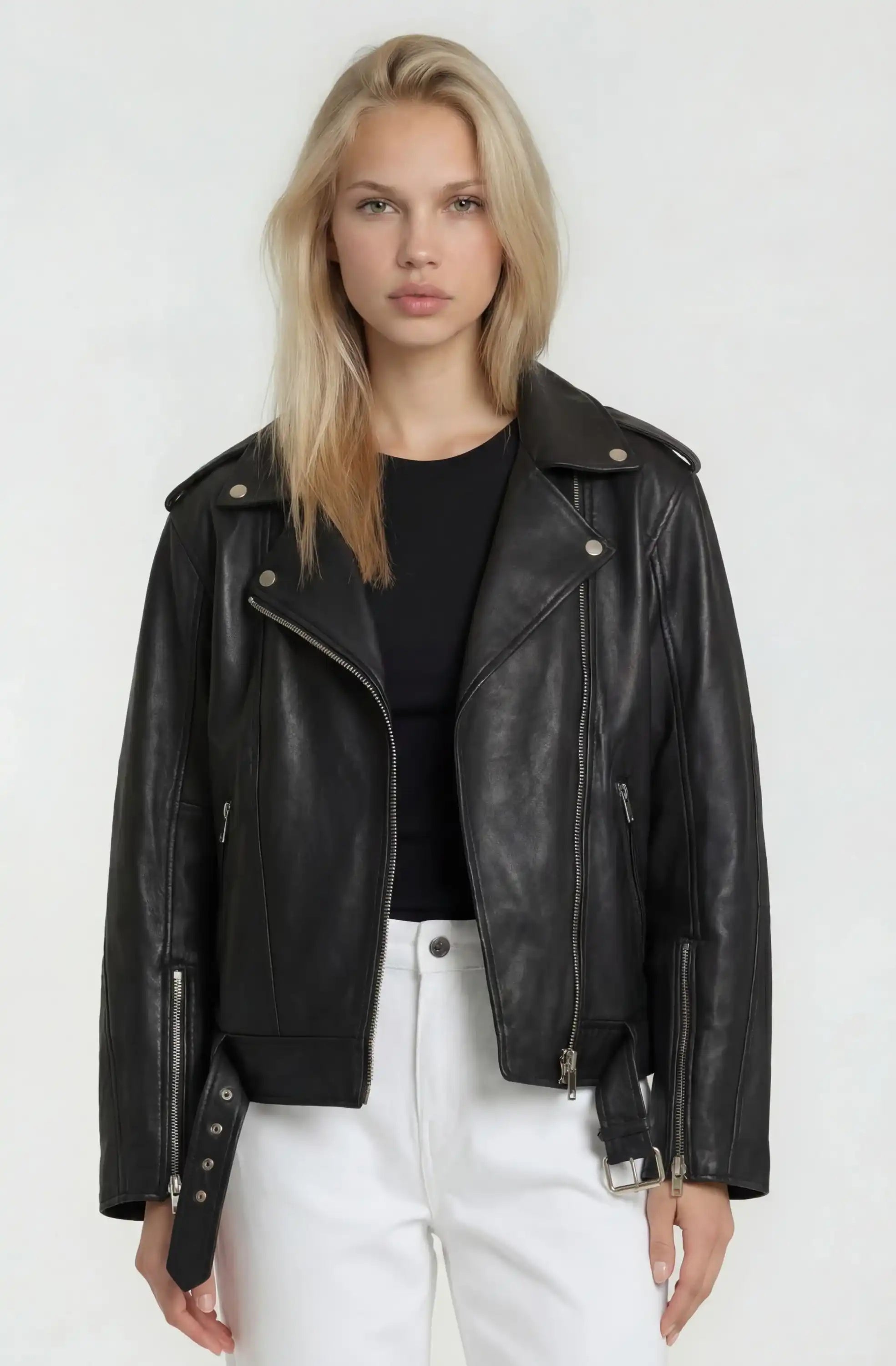 Eine Frau mit langen blonden Haaren trägt eine Lange übergroße Bikerlederjacke von trueprodigy aus weichem Schafsleder, steht vor einem schlichten hellen Hintergrund und schaut mit neutralem Blick direkt in die Kamera.