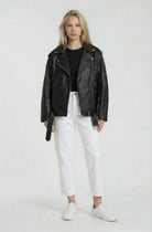 Eine Person steht vor der Kamera und trägt eine schwarze Lange übergroße Bikerlederjacke von trueprodigy, gestylt mit einem schwarzen Hemd, einer weißen Hose mit hoher Taille und weißen Turnschuhen vor einem schlichten hellen Hintergrund.