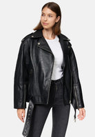 Eine Frau in einer trueprodigy Dua Lange übergroßen Bikerlederjacke, weißem T-Shirt und schwarzen Jeans steht vor einem schlichten hellen Hintergrund und schaut mit neutralem Gesichtsausdruck leicht nach rechts.