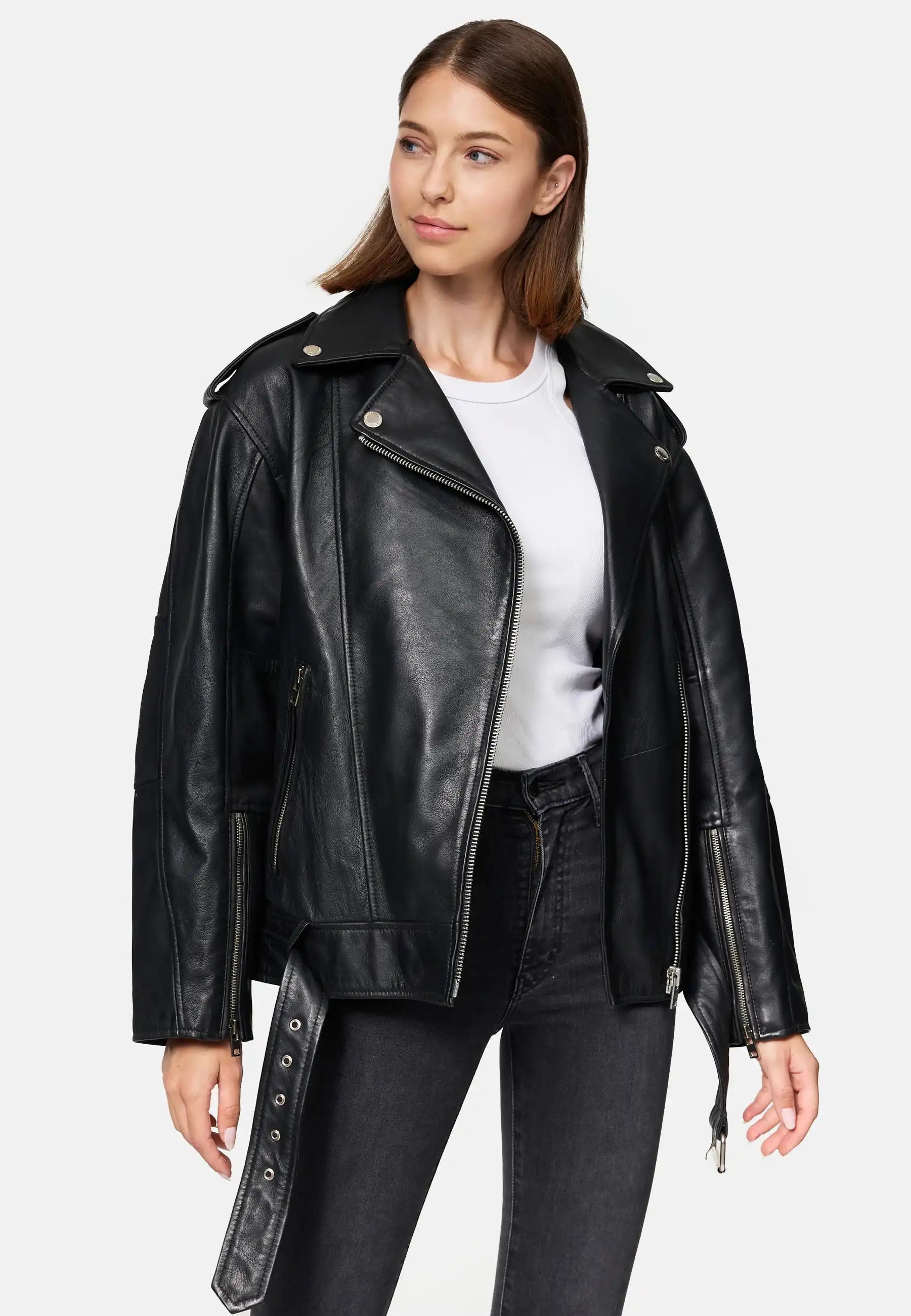 Eine Frau in einer trueprodigy Dua Lange übergroßen Bikerlederjacke, weißem T-Shirt und schwarzen Jeans steht vor einem schlichten hellen Hintergrund und schaut mit neutralem Gesichtsausdruck leicht nach rechts.