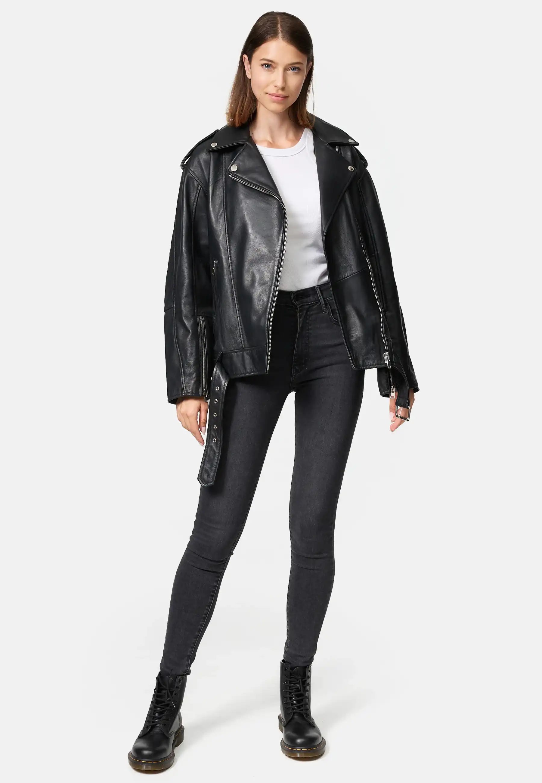 Eine Frau mit glattem braunem Haar und neutralem Gesichtsausdruck trägt eine Trueprodigy Dua Lange übergroße Bikerlederjacke, ein weißes T-Shirt, hochgekrempelte dunkle Jeans und schwarze Schnürstiefel. Sie steht vor einem schlichten hellen Hintergrund.