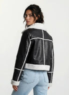 Eine Person mit langen dunklen Haaren trägt eine trueprodigy Damen-Lederjacke mit weißen Teddy-Kontrasten & asymmetrischem Reißverschluss und hellblaue Jeans, steht mit dem Rücken zur Kamera und schaut über die Schulter vor einem schlichten Hintergrund.