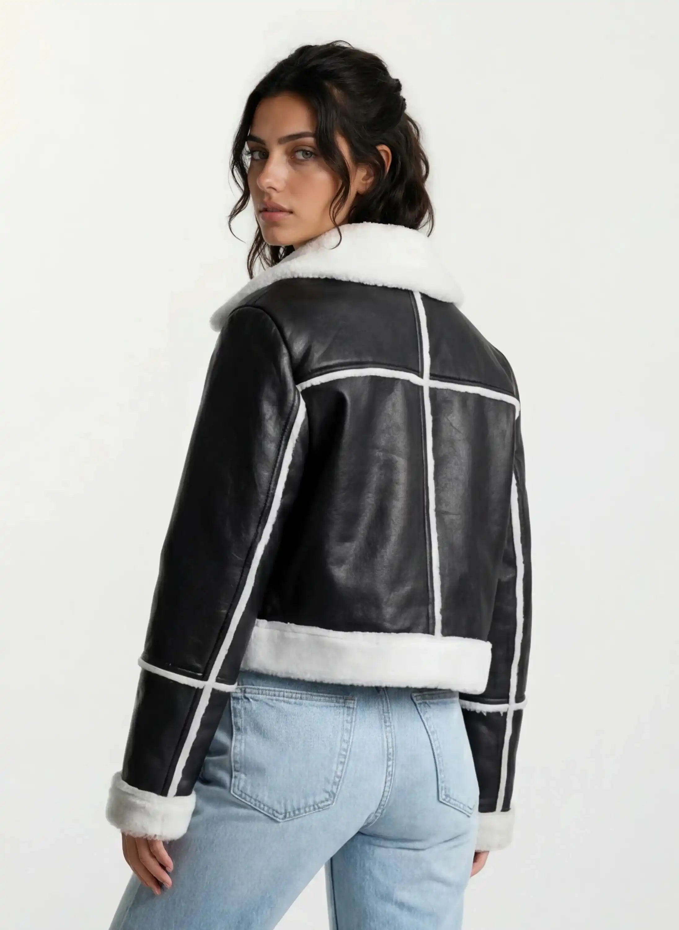 Eine Person mit langen dunklen Haaren trägt eine trueprodigy Damen-Lederjacke mit weißen Teddy-Kontrasten & asymmetrischem Reißverschluss und hellblaue Jeans, steht mit dem Rücken zur Kamera und schaut über die Schulter vor einem schlichten Hintergrund.