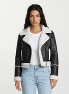 Eine Frau mit langen dunklen Haaren trägt die trueprodigy Damen-Lederjacke mit weißen Teddy-Kontrasten & asymmetrischem Reißverschluss über einem weißen T-Shirt und hellblauen Jeans, vor einem schlichten hellen Hintergrund.