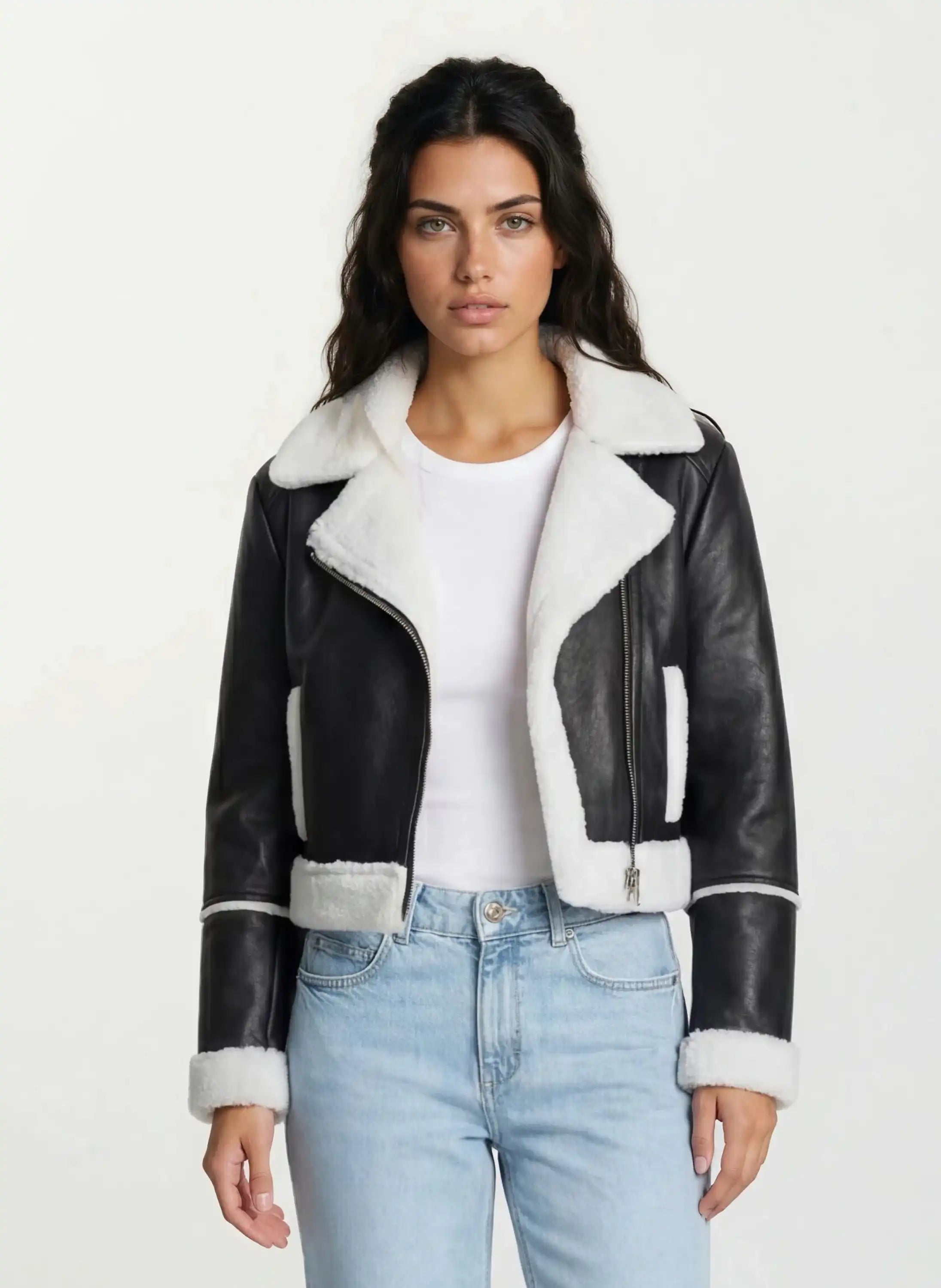 Eine Frau mit langen dunklen Haaren trägt die trueprodigy Damen-Lederjacke mit weißen Teddy-Kontrasten & asymmetrischem Reißverschluss über einem weißen T-Shirt und hellblauen Jeans, vor einem schlichten hellen Hintergrund.