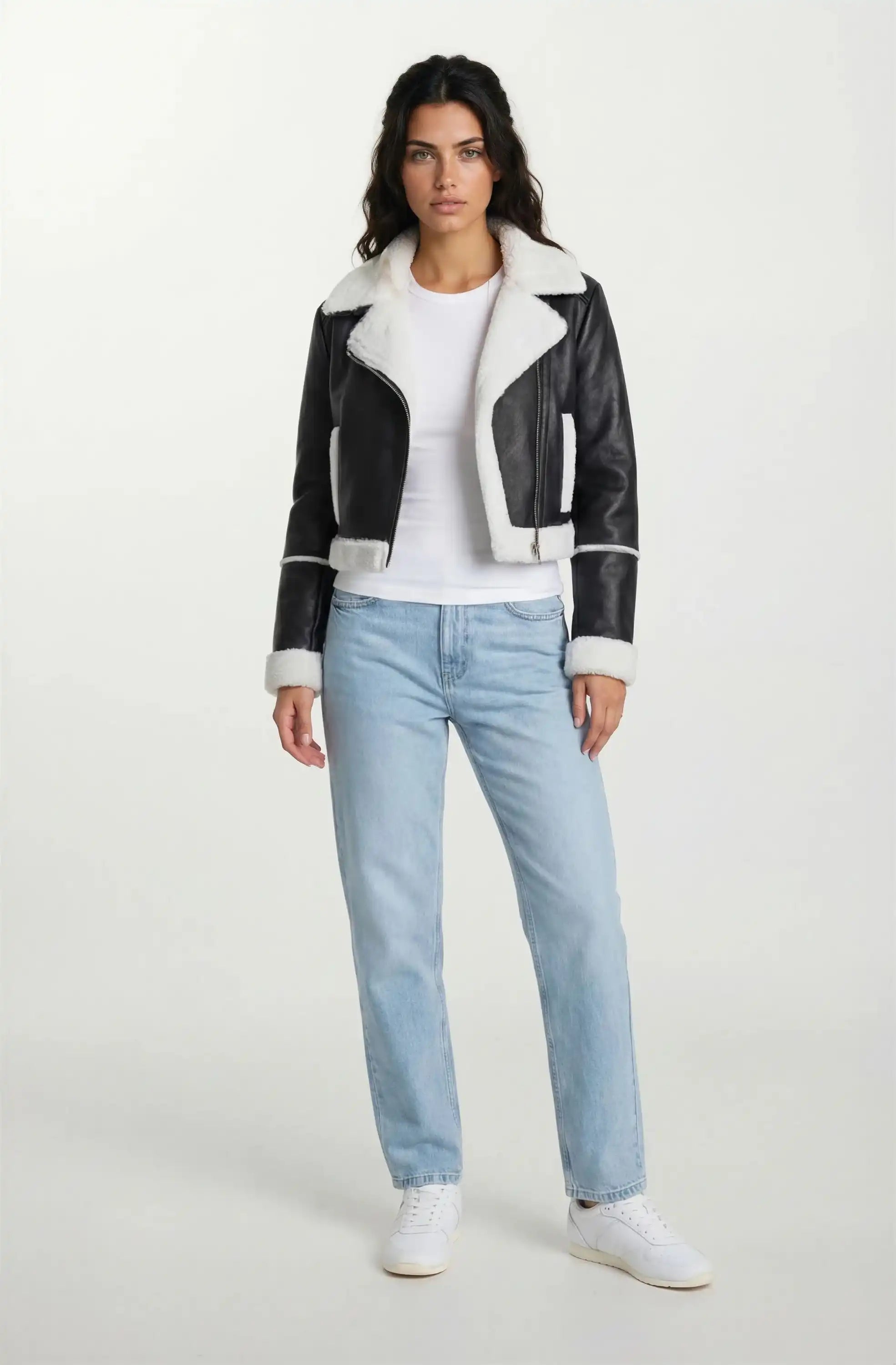 Eine Frau sieht nach vorne in einer trueprodigy Damen-Lederjacke mit weißen Teddy-Kontrasten & asymmetrischem Reißverschluss über einem weißen T-Shirt, hellblauen Jeans und weißen Turnschuhen vor einem schlichten hellen Hintergrund.