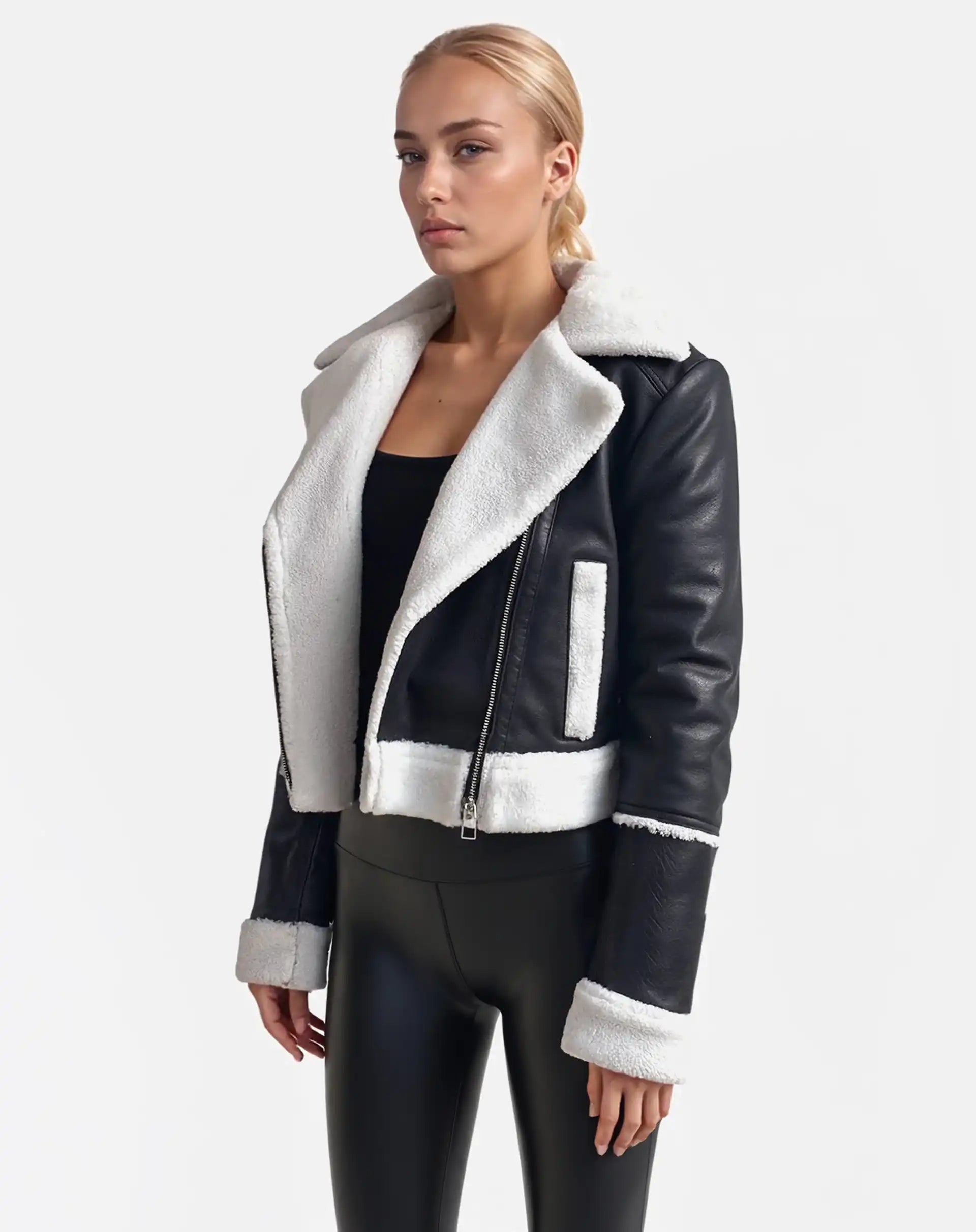 Vor weißem Hintergrund trägt eine Frau von trueprodigy eine Damen-Lederjacke mit weißen Teddy-Kontrasten und asymmetrischem Reißverschluss, dazu ein schwarzes Top und glänzende Hosen. Sie hat blonde, zurückgebundene Haare und blickt neutral.