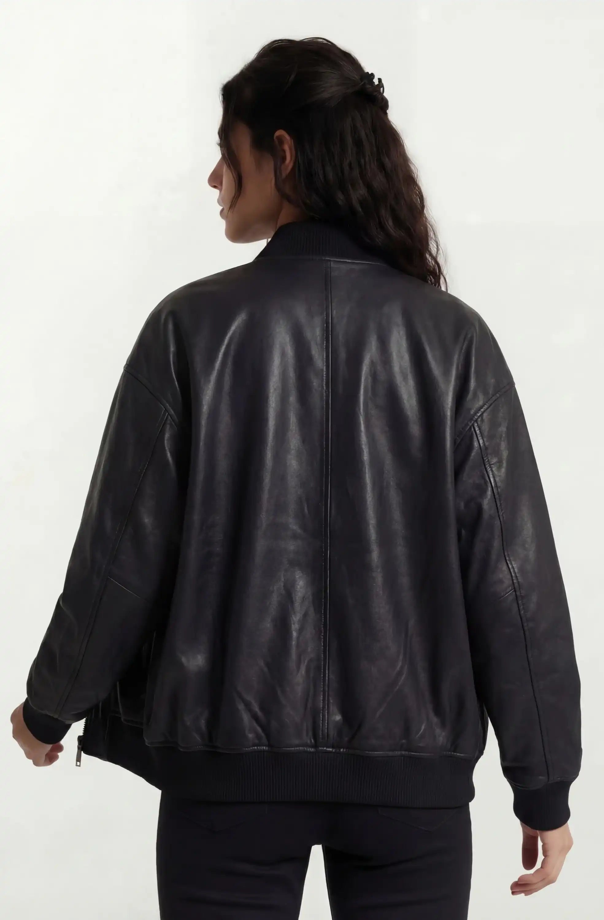 Eine Person mit langen, gewellten dunklen Haaren wendet sich von der Kamera ab und trägt eine trueprodigy Oversize-Bomberjacke aus Leder mit Rippbündchen & schwarzem Zipper und eine dunkle Hose vor einem schlichten hellen Hintergrund.